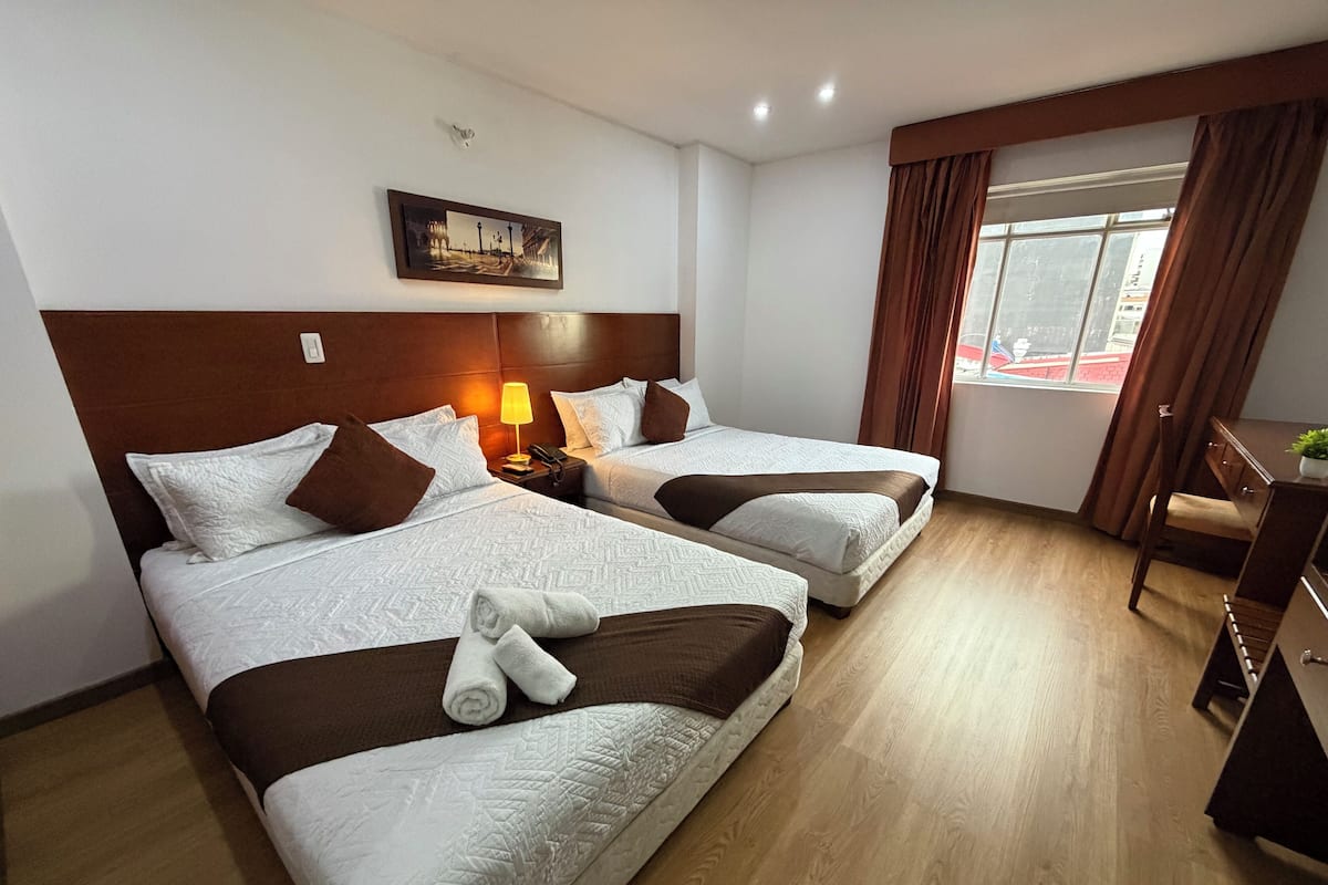 A hotel room with two beds, a desk, a lamp, a picture on the wall, and a window with curtains.