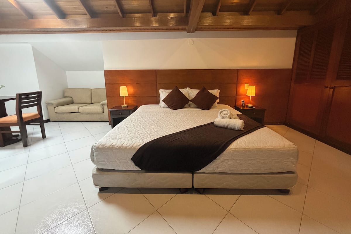 A bedroom with a bed, two bedside tables, a nightstand, a chair, a sofa, and a wooden ceiling.