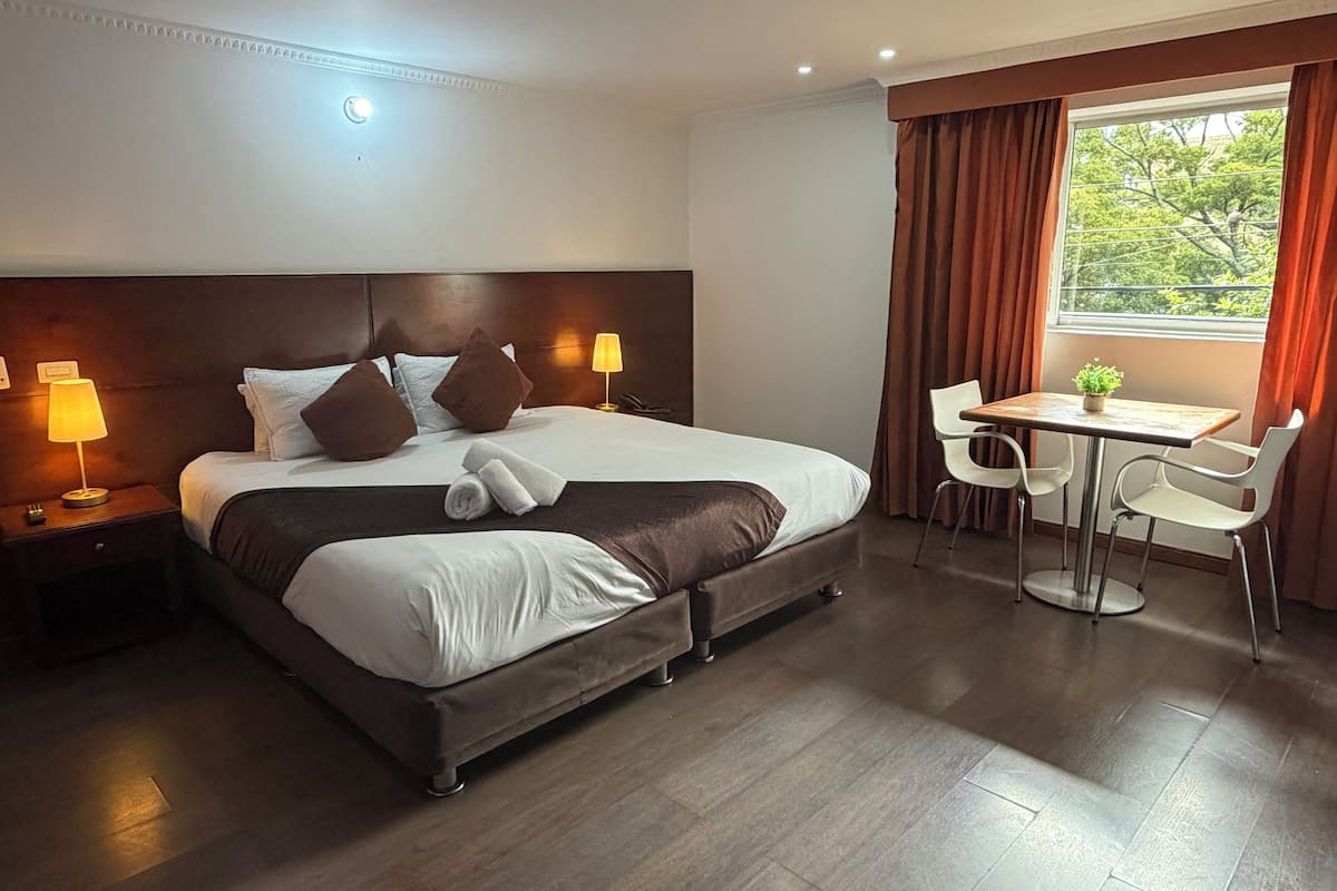 A hotel room with a bed, bedside lamps, a desk with chairs, and a window with curtains.