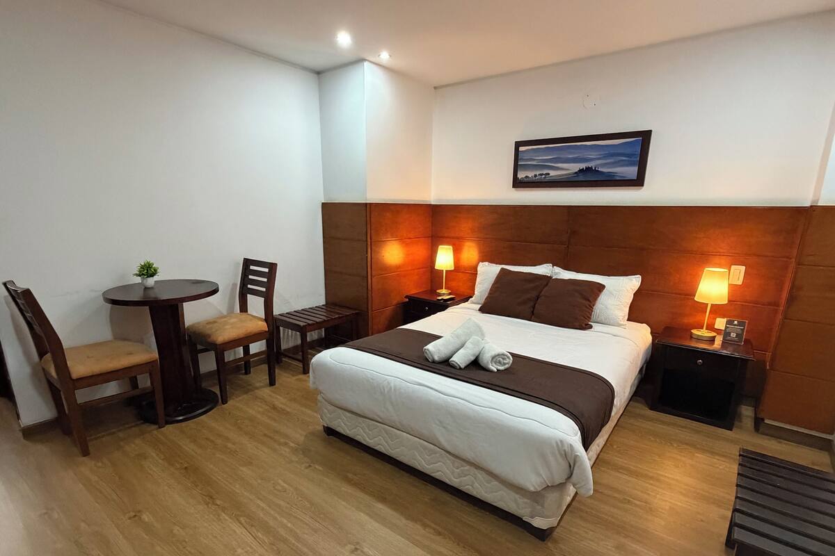 A hotel room with a bed, two bedside tables, a small round table with a chair, and a painting on the wall.