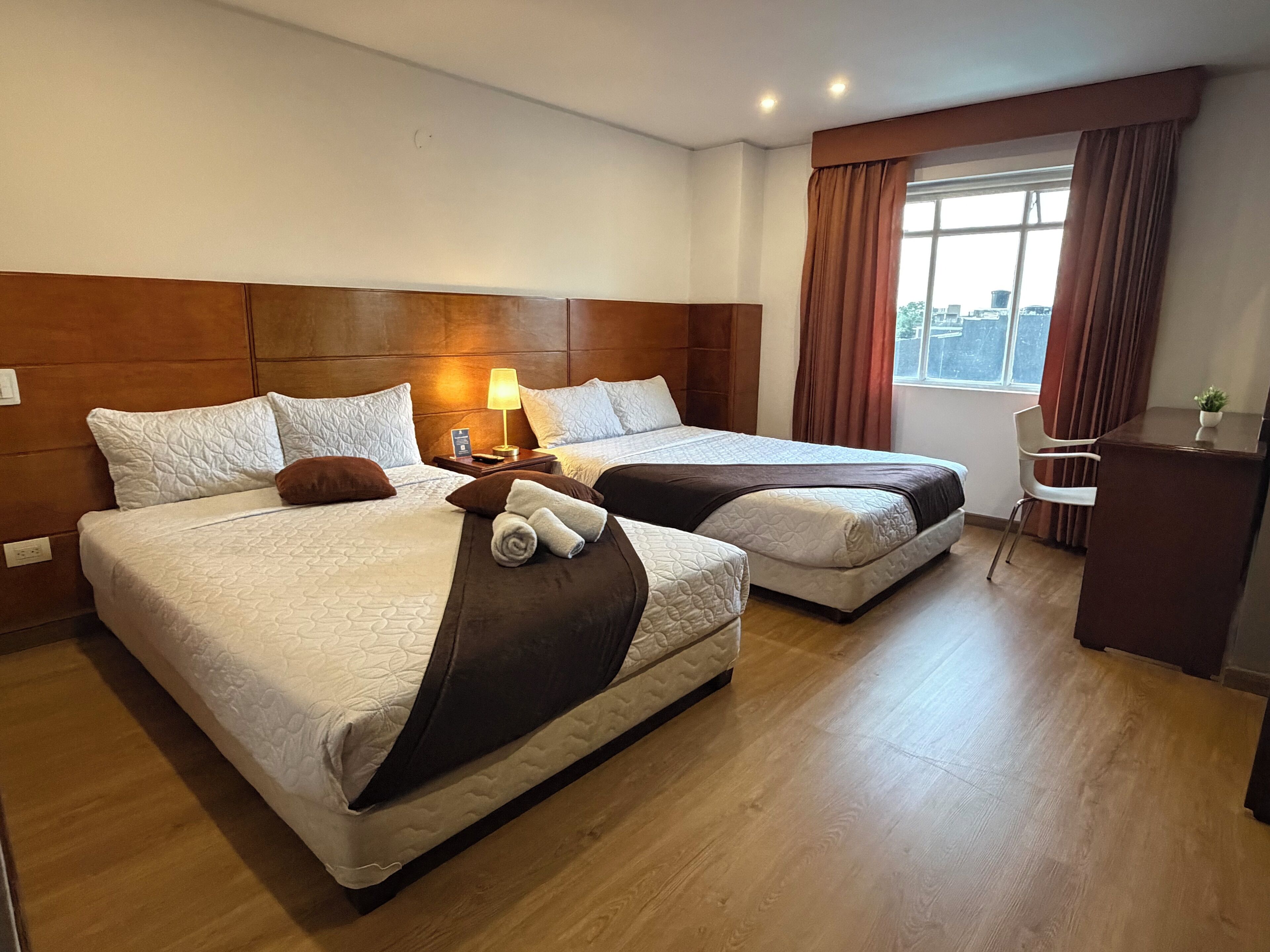 Une chambre d’hôtel avec deux lits, un bureau, une chaise, une lampe et une fenêtre avec des rideaux.