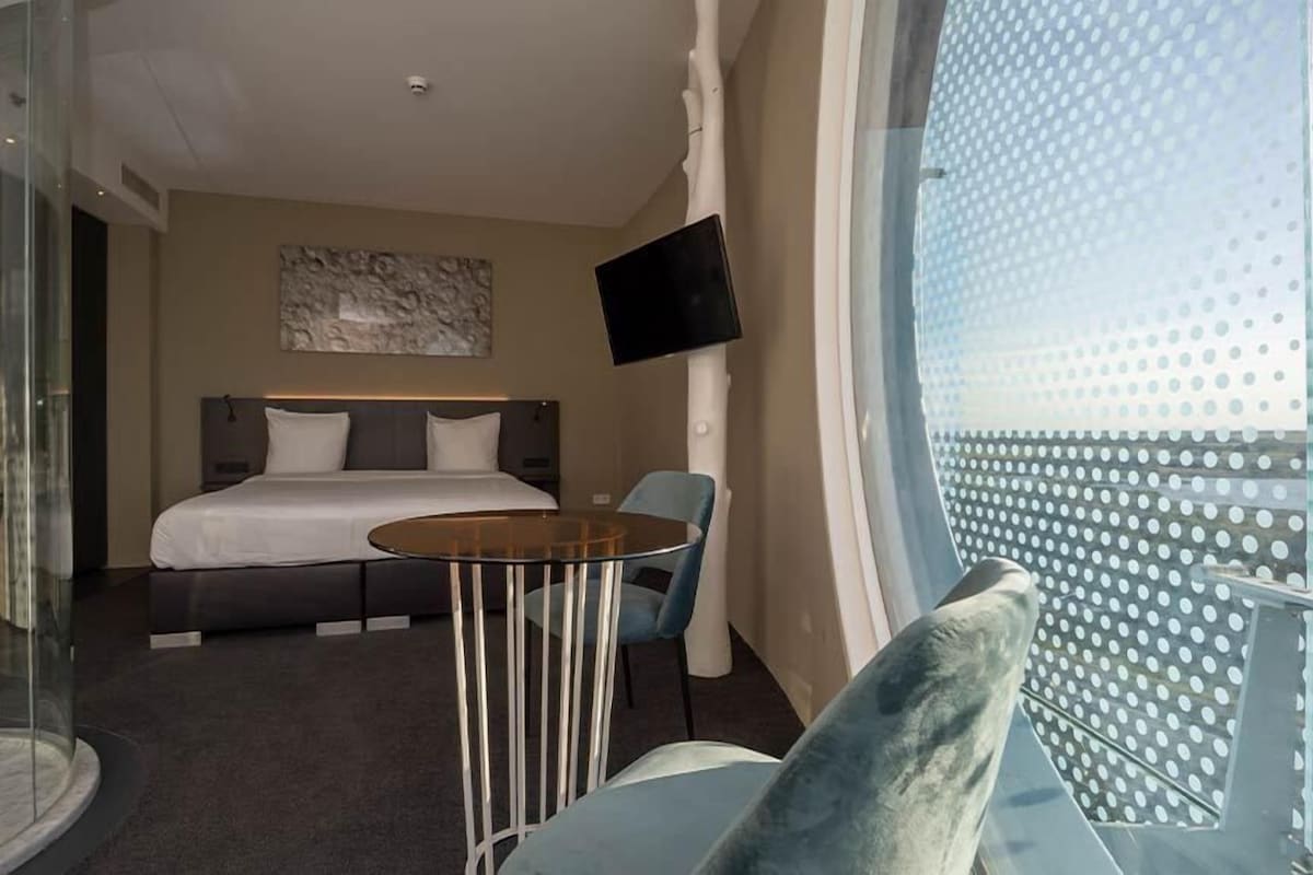 Habitación de hotel con cama, una mesita, una silla, televisión y ventana grande con vistas a la ciudad.