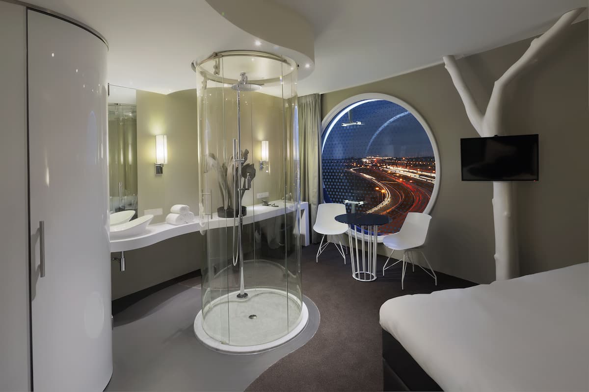 Habitación de hotel moderna con ducha de vidrio, espejo redondo, televisor grande y vistas a un paisaje urbano nocturno.
