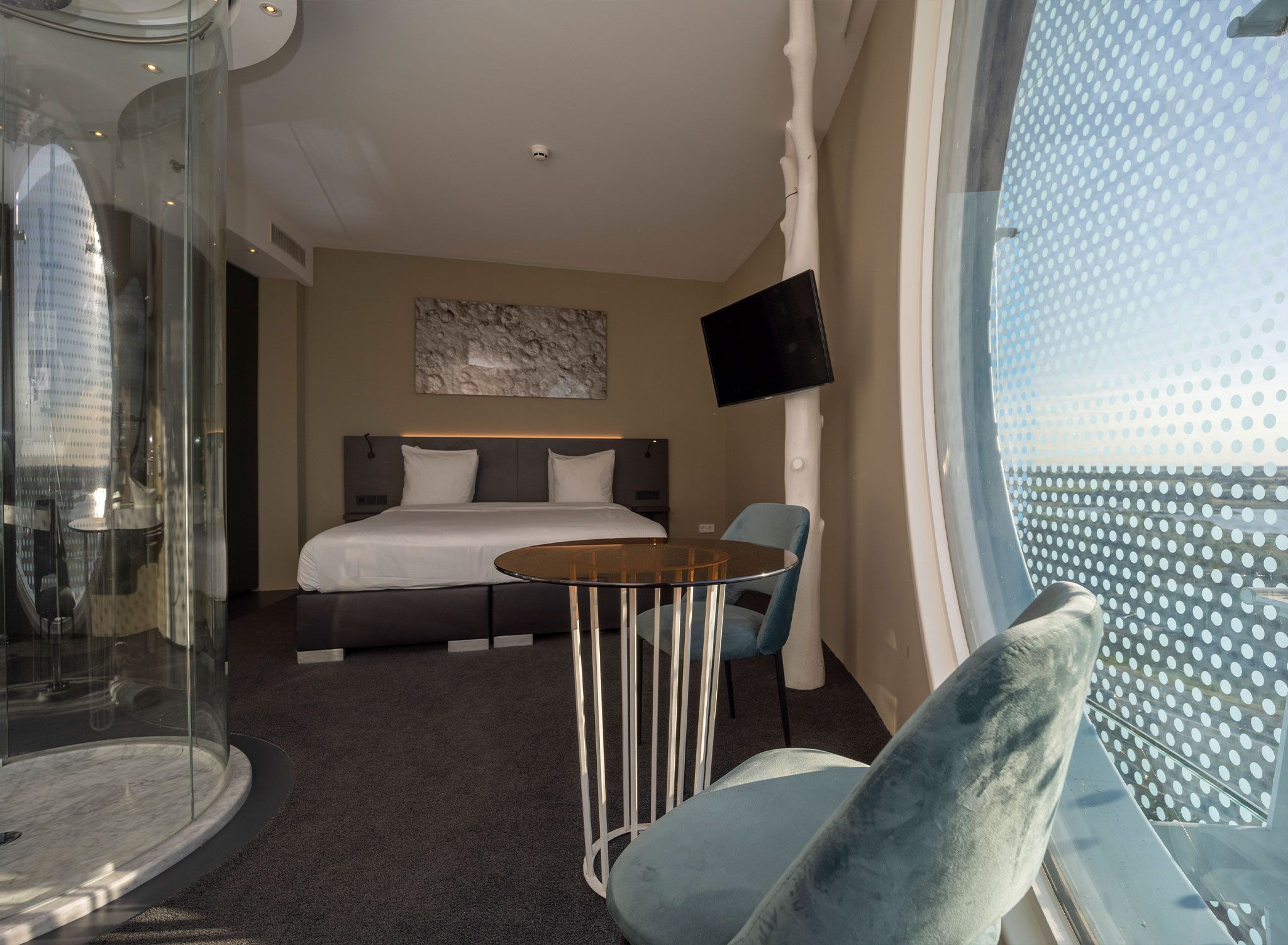 A hotel room with a bed, a glass partition, a round table, a chair, and a TV on the wall.