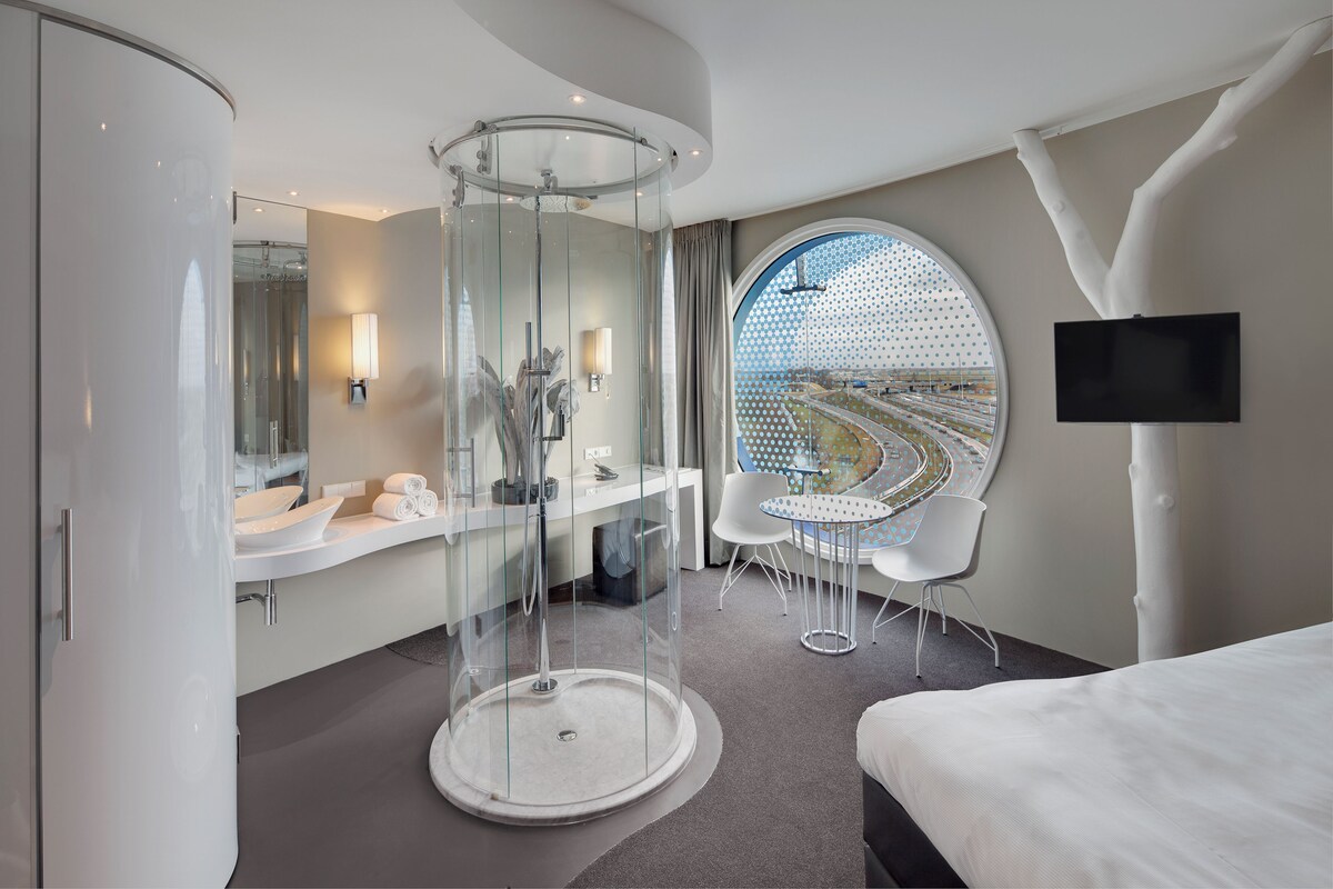 A modern hotel room with a curved shower enclosure, a round mirror, a bed, a TV mounted on the wall, and a view of a train track through a large window.
