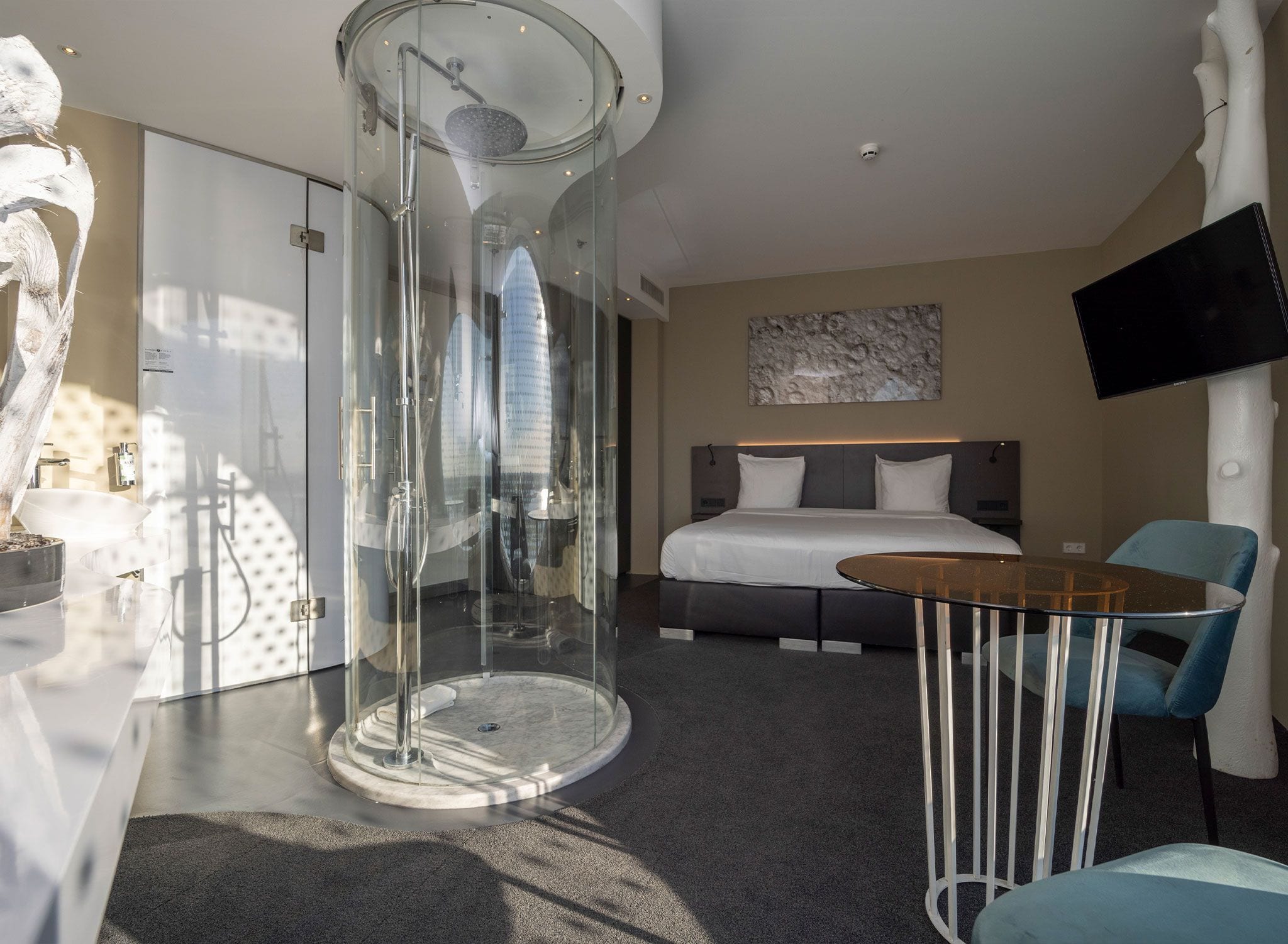A modern hotel room with a glass-enclosed shower, a bed with a headboard, a flat-screen TV mounted on the wall, and a small round table.