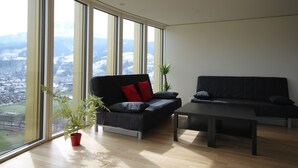 Een moderne woonkamer met een zwarte bank, rode kussens en een kleine tafel. Er staat een potplant en er is uitzicht op een besneeerd landschap door grote ramen.