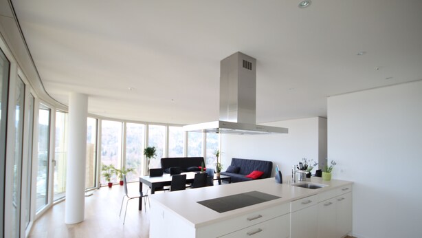 Een moderne keuken met een groot eiland, witte kasten en een rvs afzuigkap. Er is een zithoek met een zwarte tafel en stoelen, en een bank met een rode kussens. Grote ramen zorgen voor natuurlijk licht en uitzicht op het buitenleven.