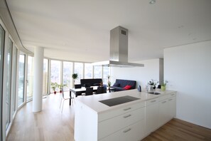 A modern kitchen with a large island, white cabinetry, and a stainless steel range hood. There is a dining area with a black table and chairs, and a sofa with a red cushion. Large windows provide natural light and a view of the outdoors. - Allmend Apartments (Lucerne)