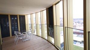 Een balkon met een houten vloer, metalen stoelen en een tafel. Er zijn grote ramen met zonwering en er is uitzicht op een stadsbeeld.
