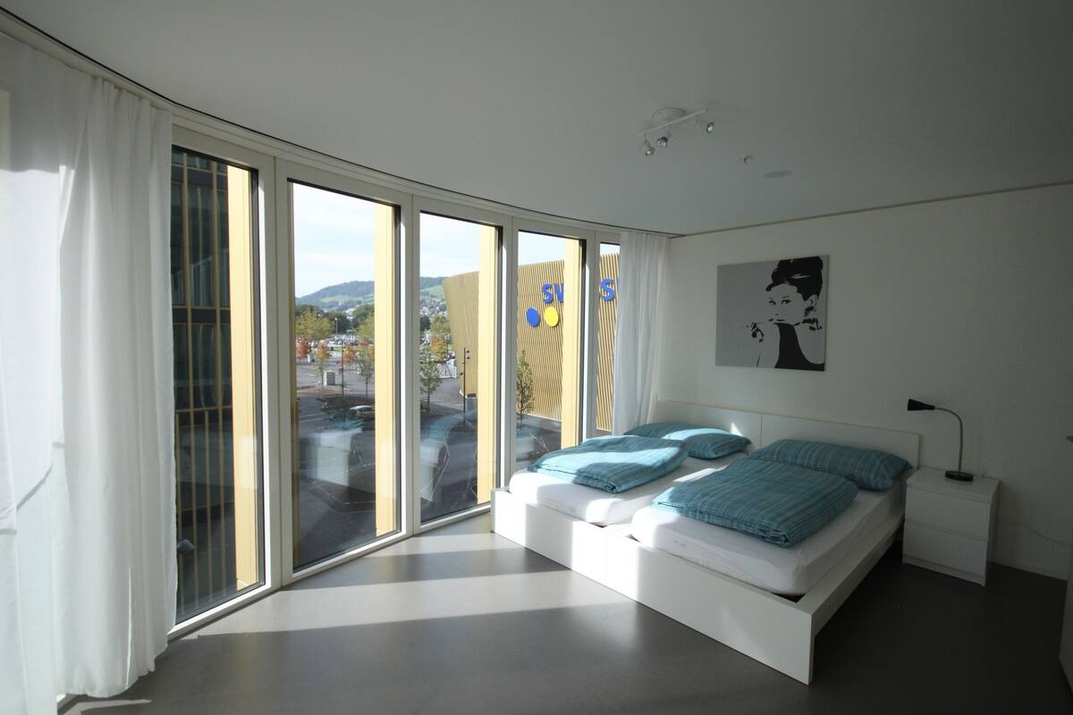 Un dormitorio moderno con un amplio ventanal corredera, una cama con ropa de cama azul, una mesita de noche y una obra de arte colgada en la pared.