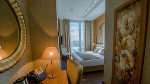 A hotel room with a bed, a desk with a telephone and a kettle, a chair, a painting, and a view of the city through the window.