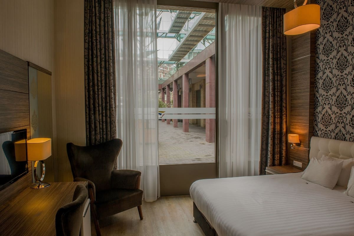 A hotel room with a bed, a chair, a desk with a lamp, a TV, and a view of an outdoor area with a glass ceiling.