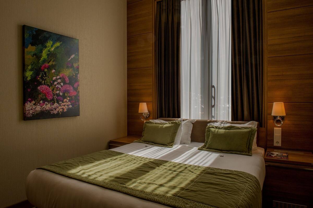 A hotel room with a large bed, two bedside lamps, a painting on the wall, and a window with curtains.