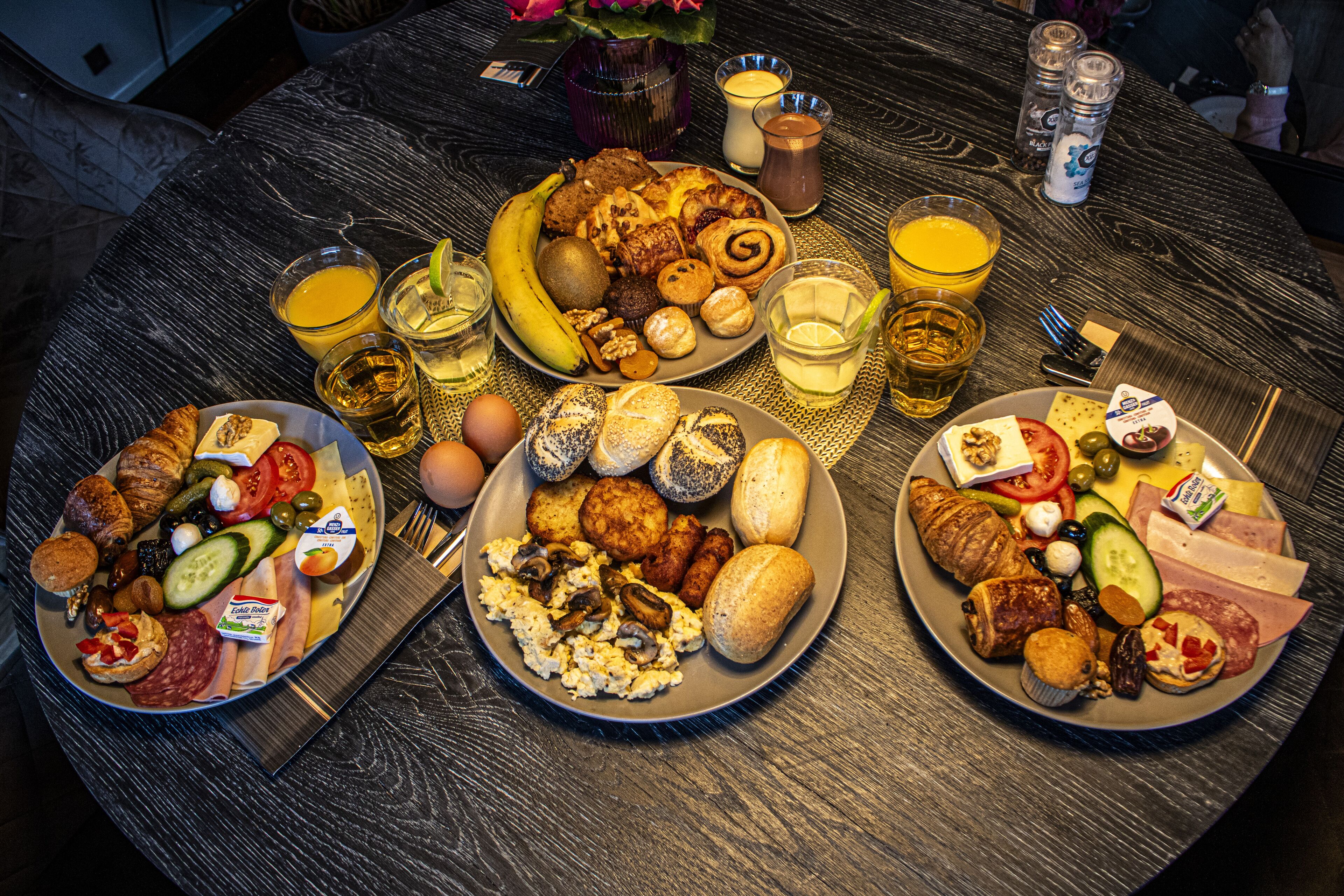 A round wooden table with three plates of assorted food, glasses of beverages, and condiments.