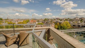 A rooftop terrace with wicker furniture, a glass balustrade, and a view of a cityscape with buildings and trees. - Grand Hotel Downtown (Amsterdam)