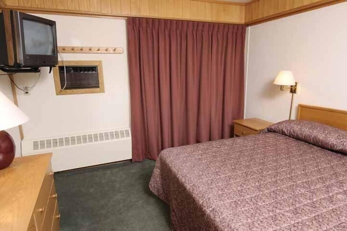 A hotel room with a bed, a television mounted on the wall, a window with curtains, a bedside table with a lamp, and a radiator.