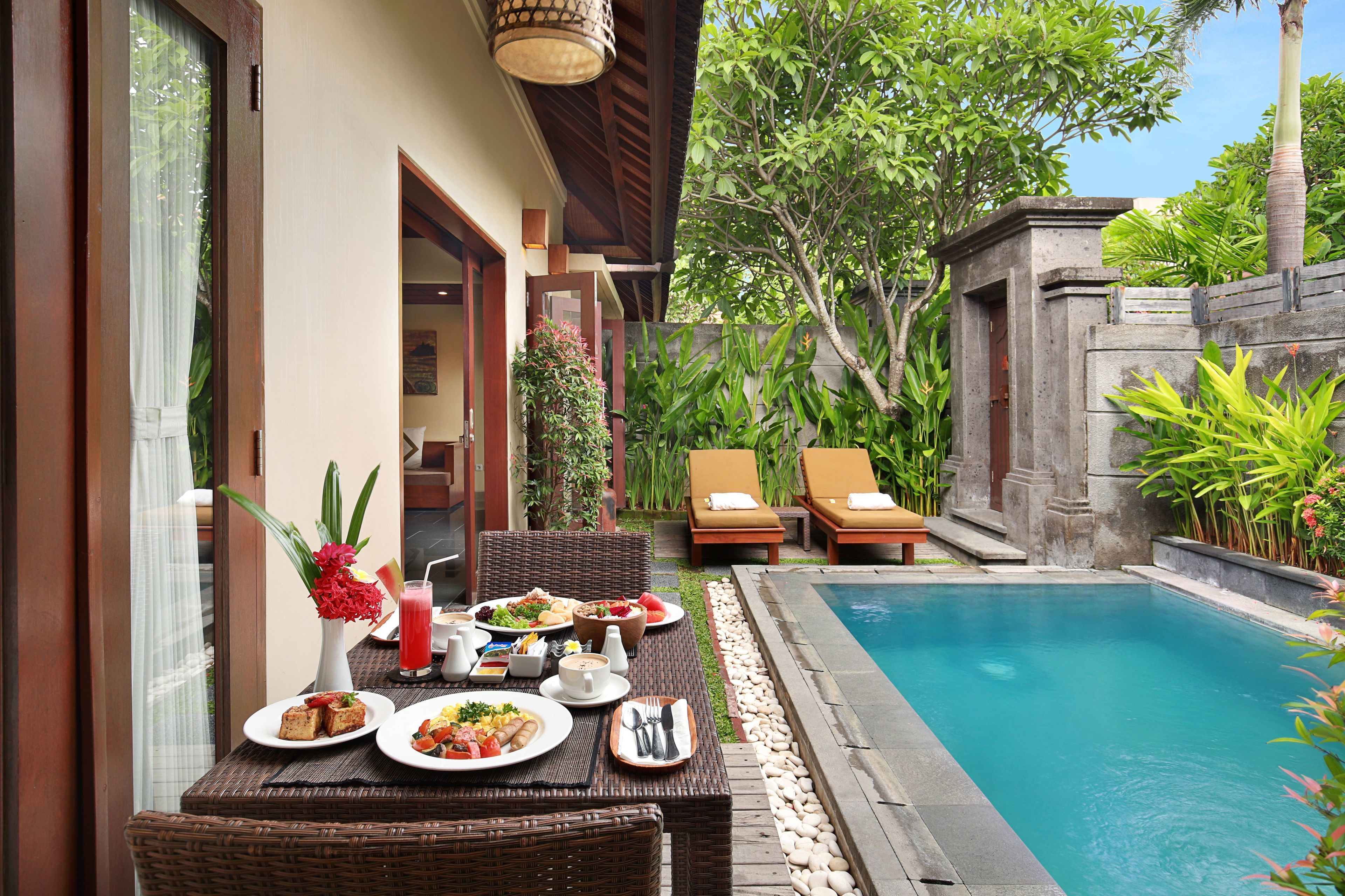 A dining area with a poolside view, featuring a table set with plates, a vase with flowers, and a drink.