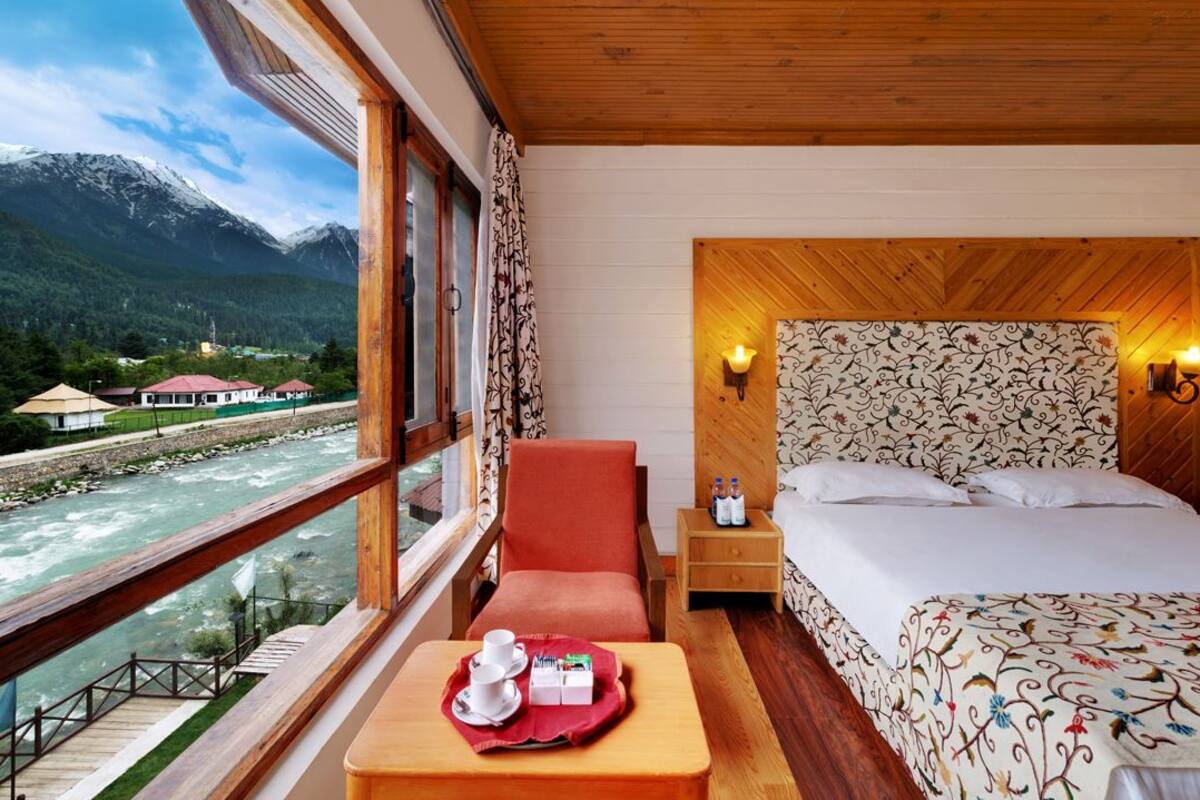 Habitación de hotel con un ventanal grande que da a un río y montañas, una cama con ropa de cama estampada, cabecero de madera y una mesita con alimentos para el desayuno.