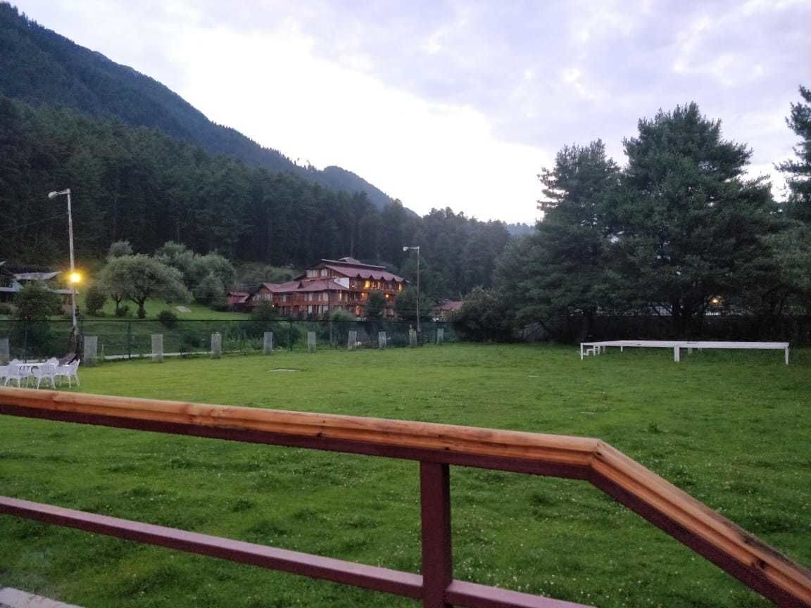 A grassy field with a wooden fence in the foreground, a building with a red roof in the background, and a mountain range.