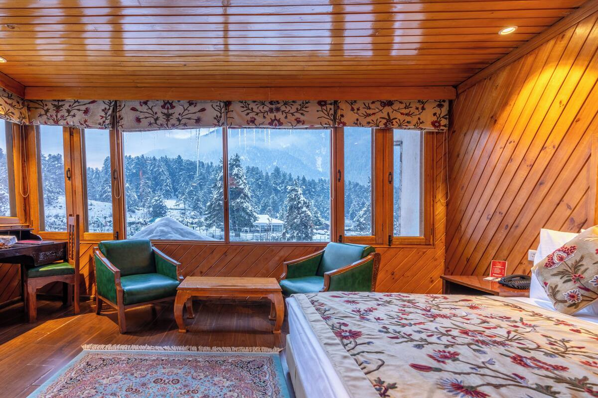 Un cuarto acogedor con una cama, dos sillas verdes, un escritorio de madera y vistas a árboles cubiertos de nieve a través de amplios ventanales.
