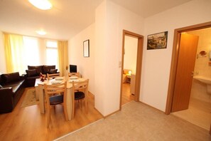 Una sala de estar moderna con un sofá marrón oscuro, una mesa de comedor de madera con sillas, y una cocina visible a través de una puerta abierta.