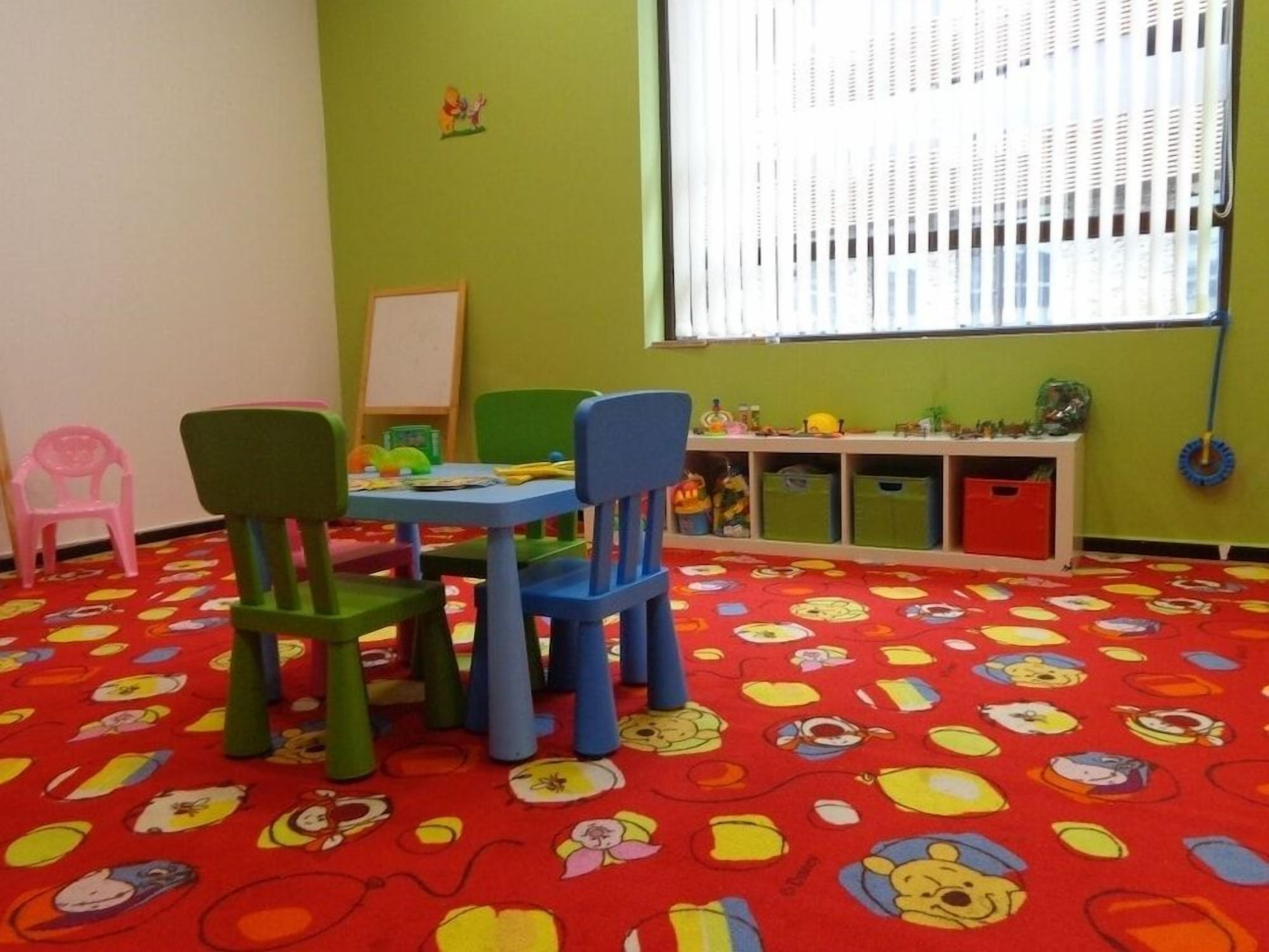 Habitación con alfombra roja que presenta personajes de caricatura, sillas verdes y azules, una mesita y una ventana con persianas.