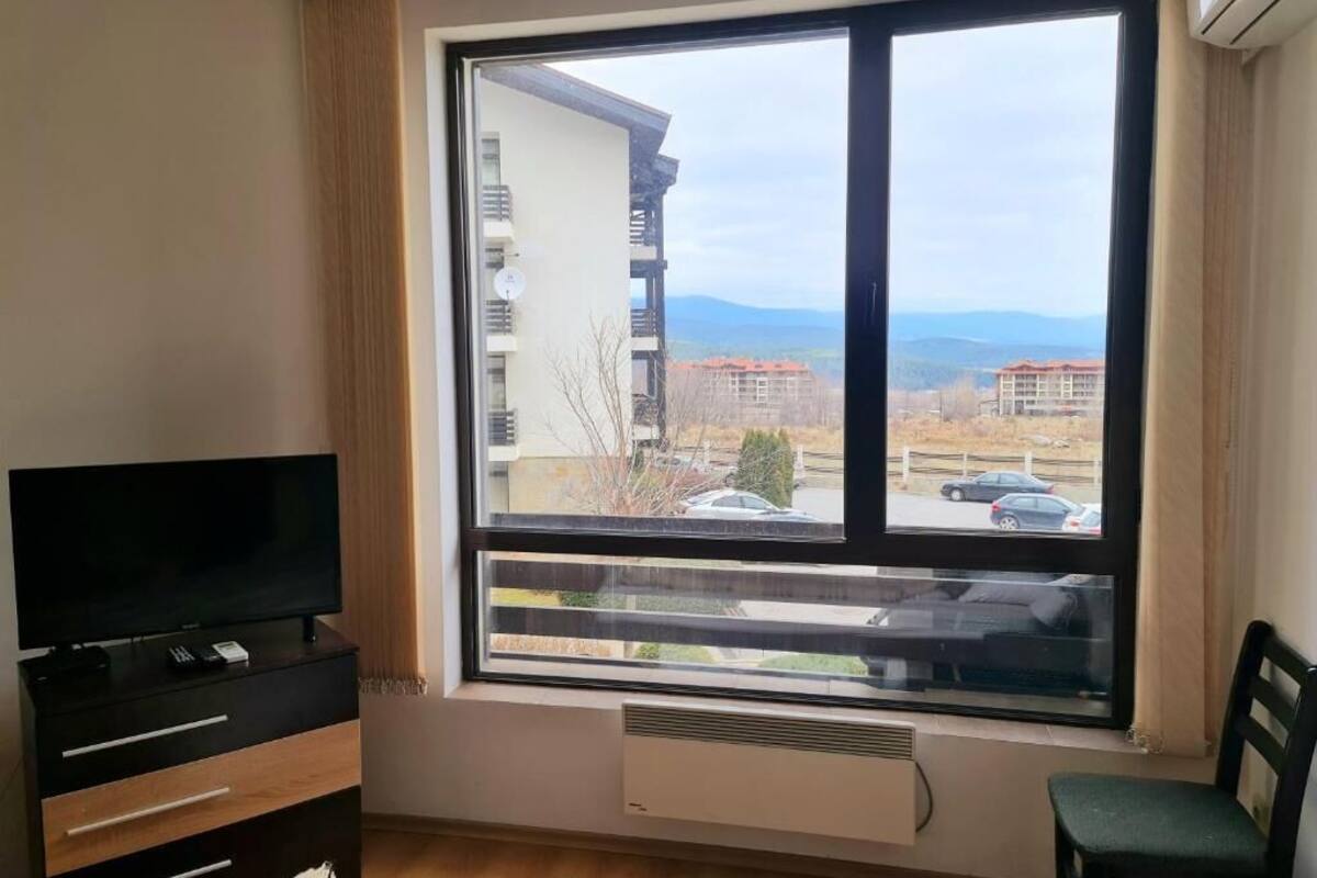 A room with a large window, a TV on a wooden cabinet, and a chair.