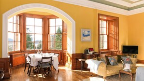 A room with a dining table set for four, a sofa with floral cushions, and large windows with wooden frames.