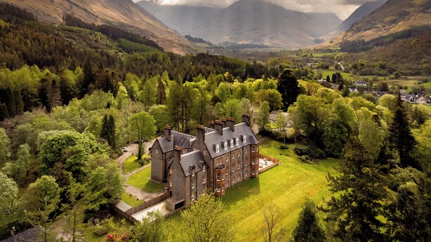 A large, historic building with multiple chimneys, surrounded by lush green fields and dense forests, with mountains in the background.