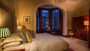 A large bed with multiple pillows, a nightstand with a lamp, a desk with a chair, a window with a view of mountains, and a chandelier.