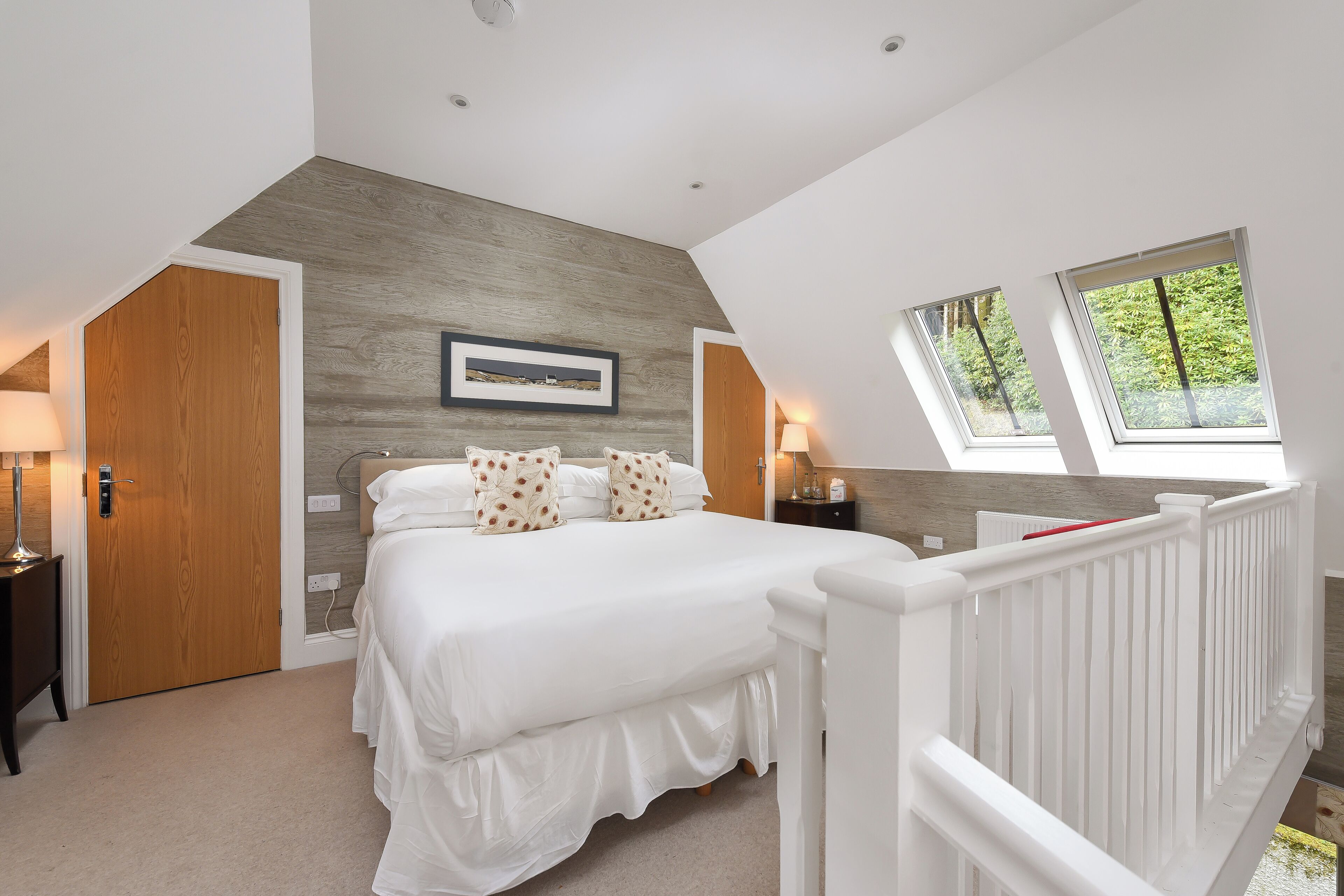 A bedroom with a large bed, a wooden accent wall, two bedside lamps, and a window with a view of greenery.