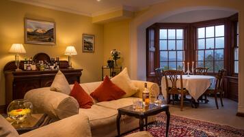 A cozy living room with a sofa, a coffee table, and a dining table set for four. There are lamps, a vase with flowers, and a view through large windows.