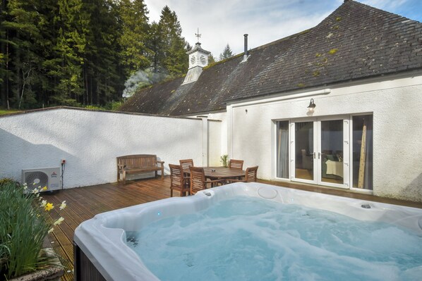 Un jacuzzi en un patio trasero con terraza de madera, una mesa de comedor con sillas y un edificio blanco con chimenea.