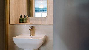 Baño con lavamanos blanco, grifo dorado y un espejo encima. Hay dos botellas en el estante sobre el espejo.