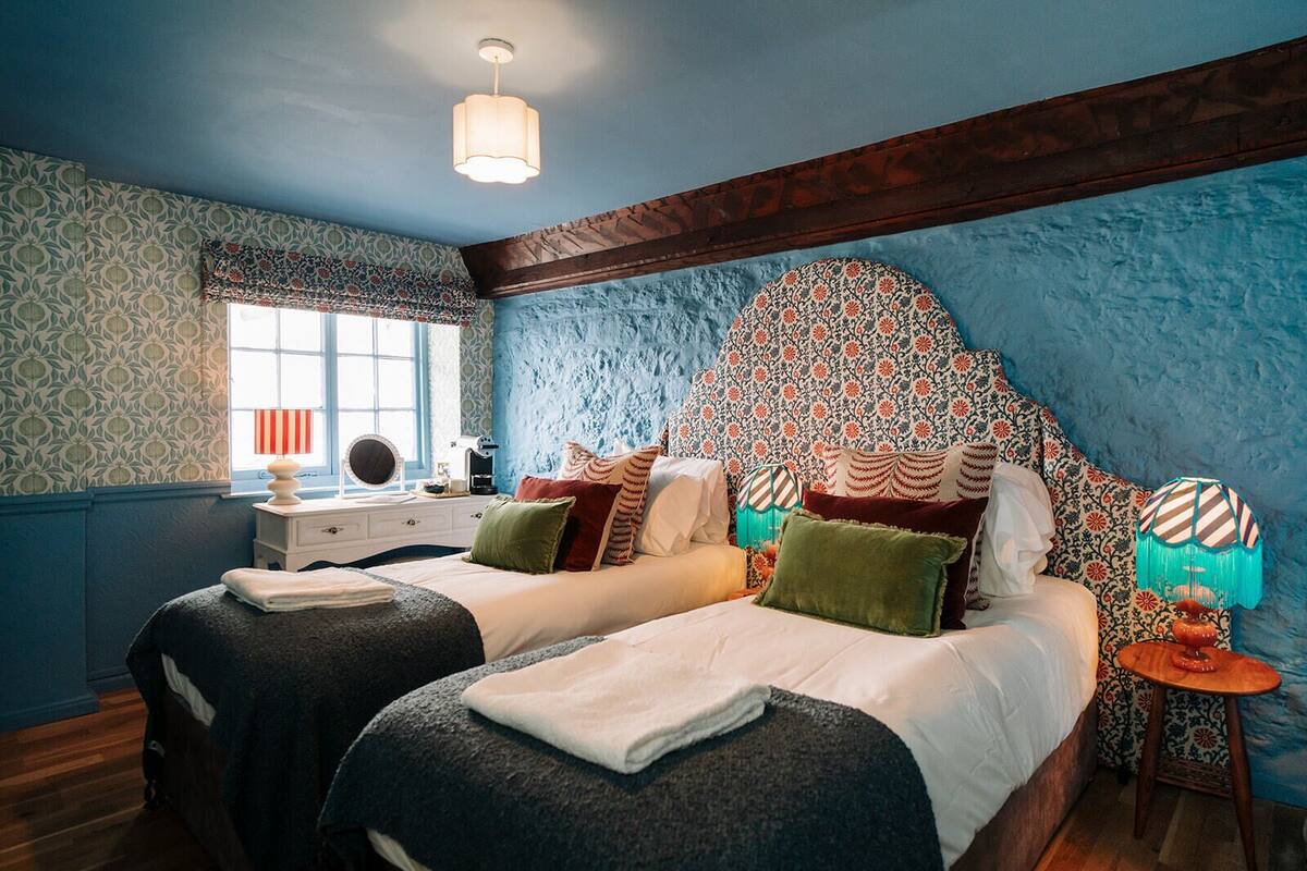 A bedroom with two beds, a window with a striped curtain, a bedside table with a lamp, and a patterned headboard.