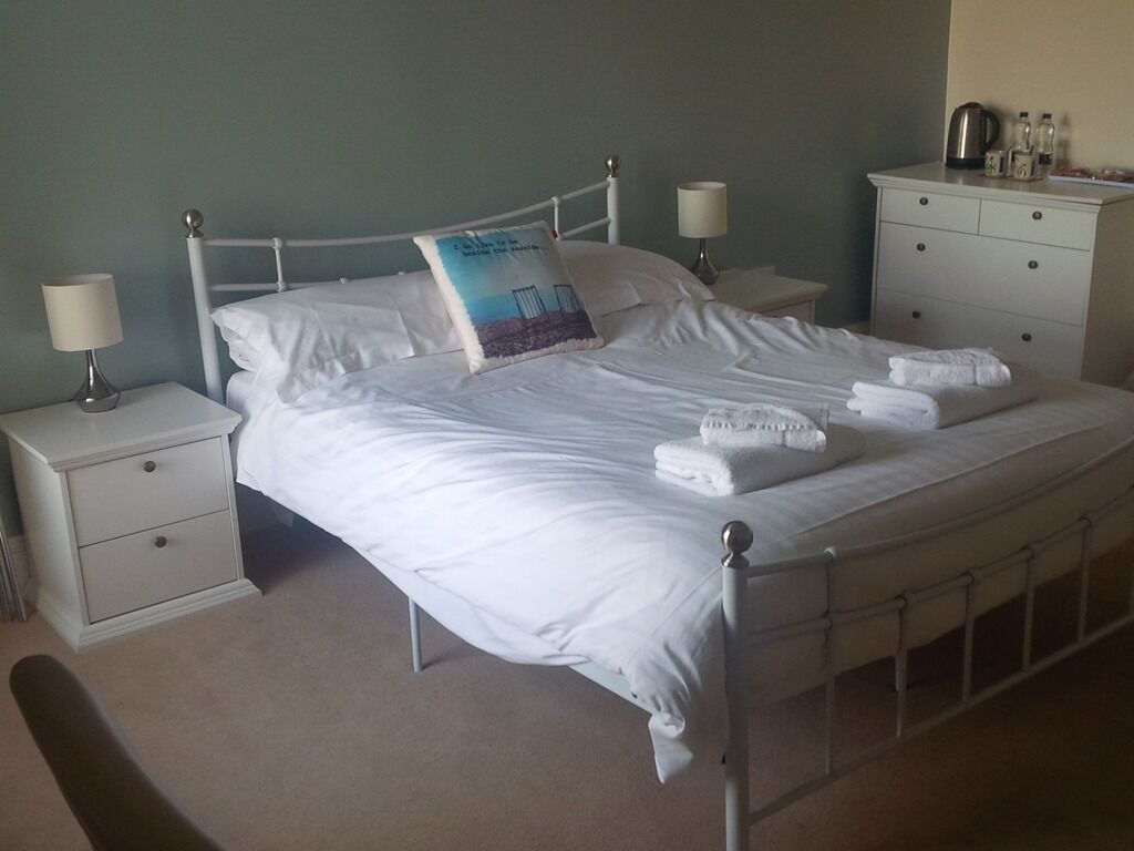 A neatly made bed with white linens and a decorative pillow, two bedside tables with lamps, a dresser with toiletries, and a beige carpeted floor.