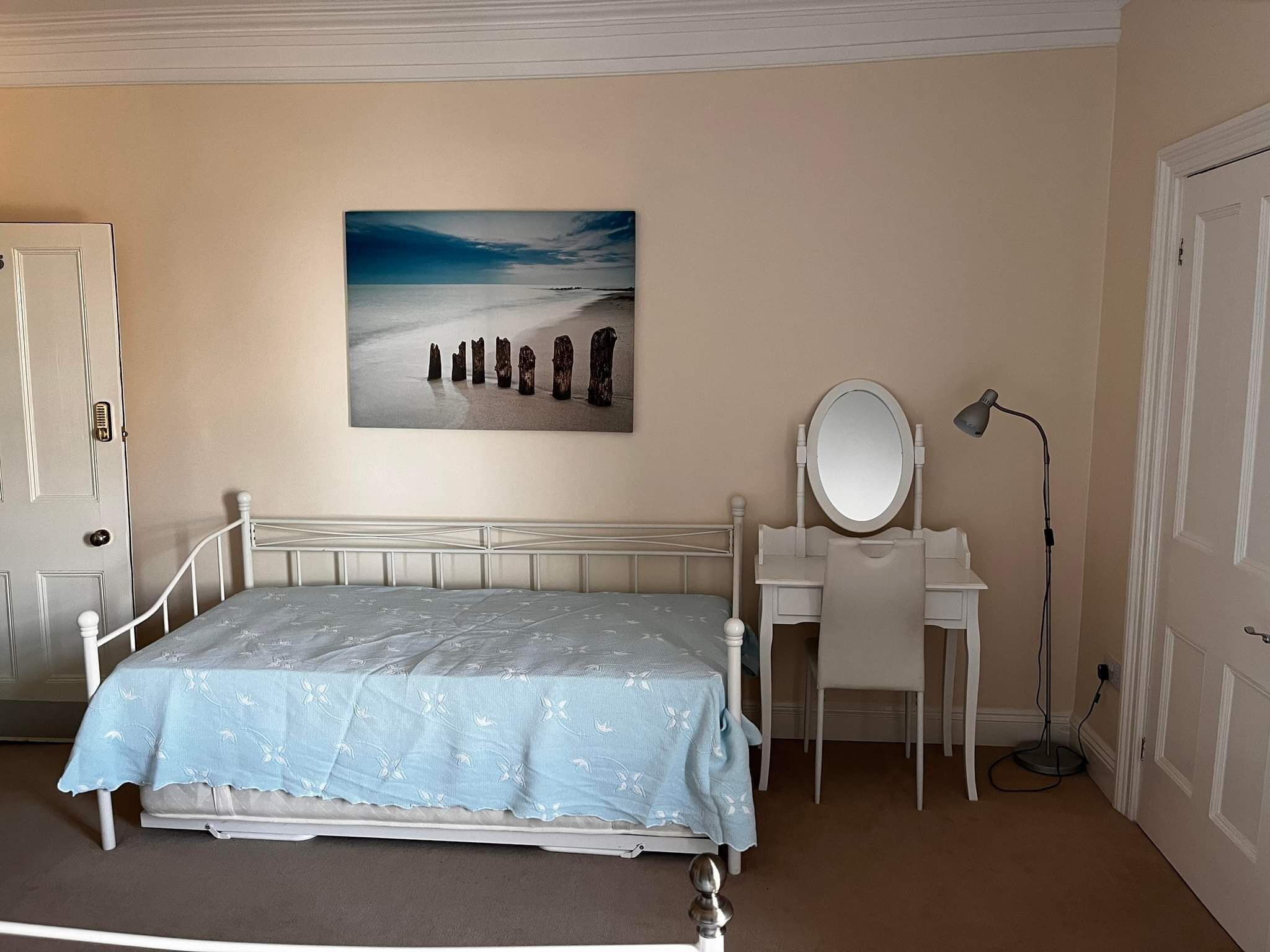Et soveværelse med seng, skrivebord med spejl, lampe og et billede på væggen.