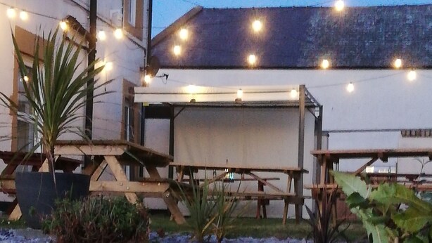 Un’area patio all’aperto con pavimento in pietra, tavoli in legno e luci a tendina.