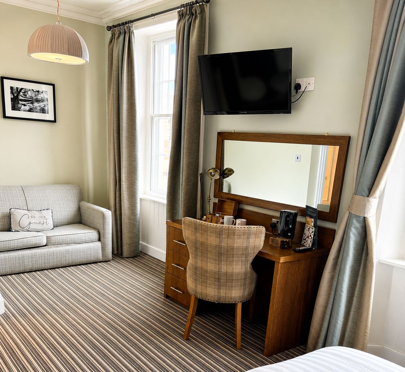 Habitación de hotel con sofá, escritorio y silla, televisor, espejo y ventana con cortinas.
