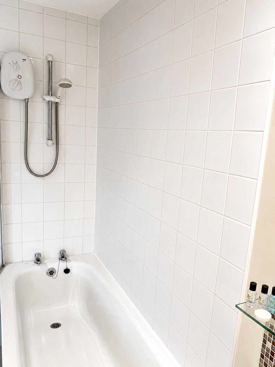 Un bagno con pareti piastrellate di bianco, una vasca da bagno, una doccia con soffione a mano e uno scaffale in vetro con articoli da toilette.