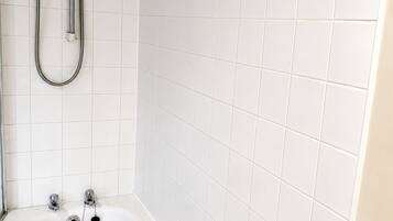 Un bagno con pareti piastrellate di bianco, una vasca da bagno, una doccia con soffione a mano e uno scaffale in vetro con articoli da toilette.