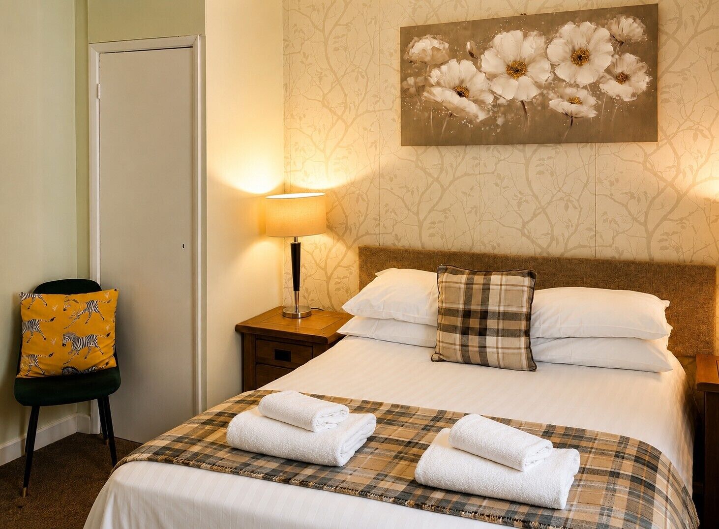 A hotel room with a bed, a nightstand with a lamp, a chair with a zebra-patterned cushion, and a wall with a floral painting.