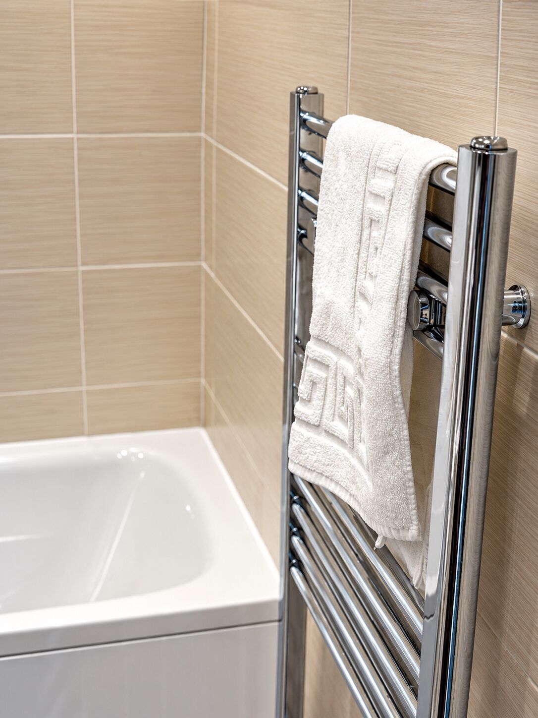 Une salle de bain avec une baignoire blanche, un porte-serviettes chromé sur lequel est posée une serviette blanche, et des murs recouverts de carreaux beiges.