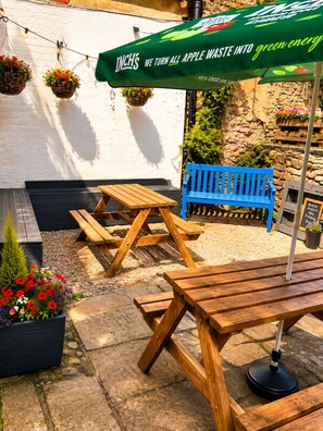 A patio area with wooden benches and picnic tables, a blue bench, hanging flower baskets, and a green umbrella with a slogan. - The Castle Tavern (Richmond)