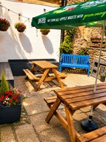 A patio area with wooden benches and picnic tables, a blue bench, hanging flower baskets, and a green umbrella with a slogan.