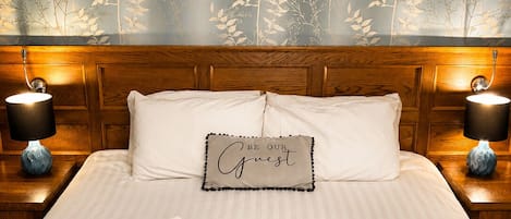A neatly made bed with a sign that reads 'BE OUR Guest,' two bedside lamps, and a plaid-patterned area rug.