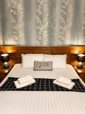 A neatly made bed with a sign that reads 'BE OUR Guest,' two bedside lamps, and a plaid-patterned area rug. - The Castle Tavern (Richmond)