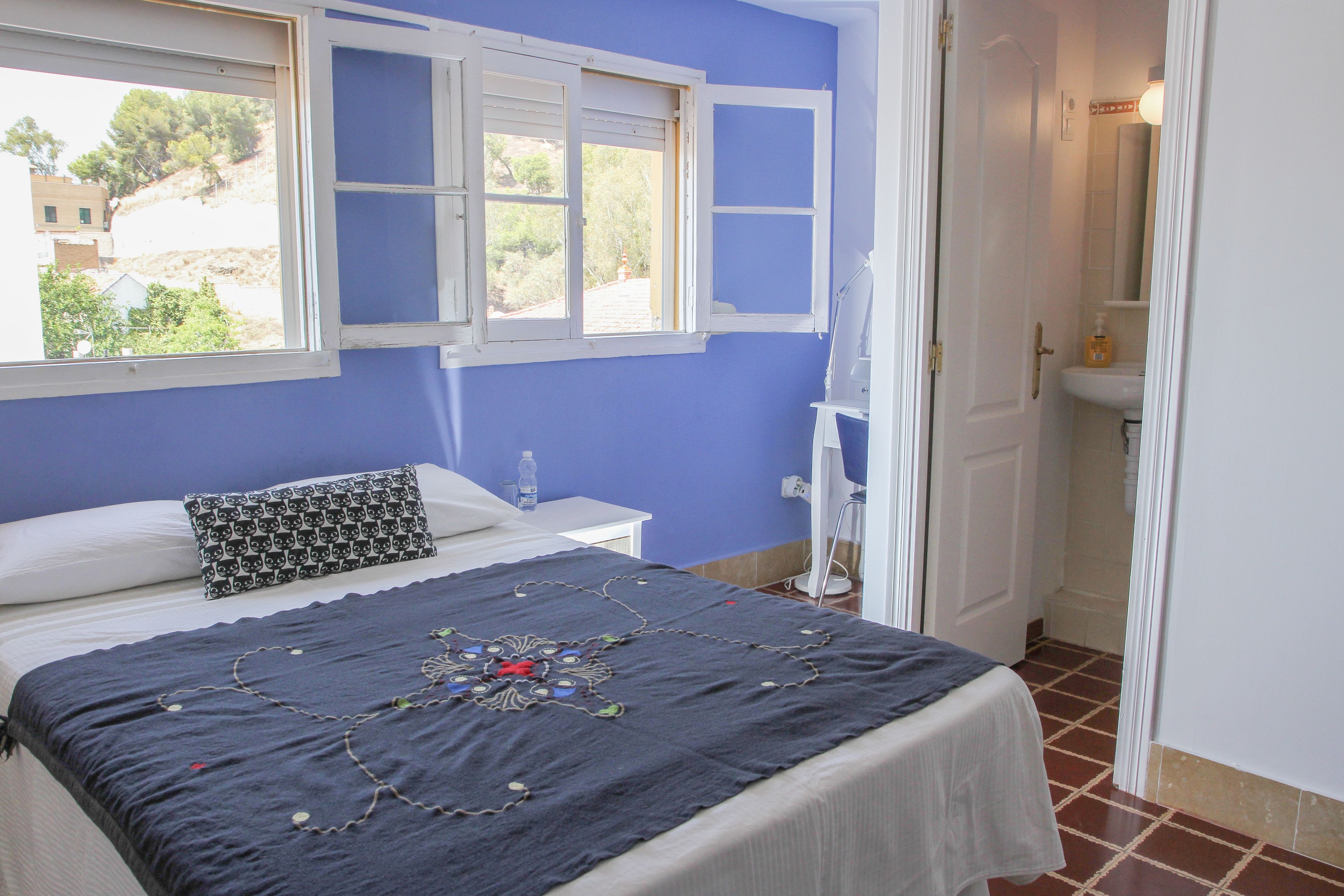 A bedroom with a bed, a window, a bedside table, and a bathroom visible through an open door.