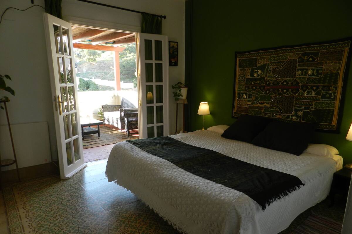 Une chambre à coucher comprenant un lit, une table de chevet, une lampe, un mur vert et une porte donnant sur un espace extérieur.